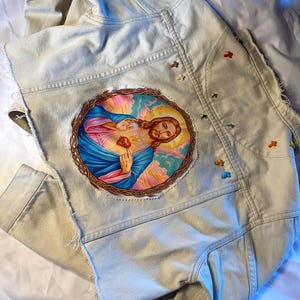 Puede incluir: Chaqueta vaquera beige claro con un parche circular que muestra una imagen religiosa. El parche representa una figura con un corazón rojo, rodeada por un halo colorido. Pequeños acentos decorativos coloridos están cosidos en la chaqueta.