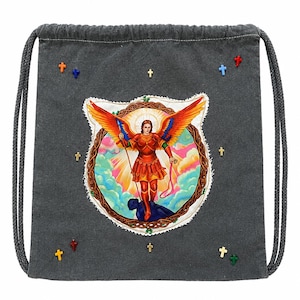 Puede incluir: Una mochila de cordón gris carbón con un parche circular central que representa a un ángel con alas naranjas y rojas sobre una figura oscura. La bolsa está decorada con pequeños adornos coloridos en forma de cruz en rojo, azul, verde y amarillo.