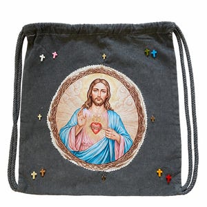 Puede incluir: Una mochila de cordón gris oscuro con un estampado circular central del Sagrado Corazón de Jesús, representado con túnica azul y rosa. La bolsa está decorada con varios pequeños dijes en forma de cruz de múltiples colores dispersos alrededor.