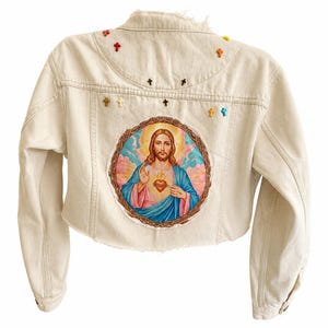 Puede incluir: Una chaqueta de mezclilla color crema con un gran parche circular en la espalda que representa una figura religiosa con un halo y un sagrado corazón. El canesú superior de la espalda está decorado con pequeños adornos en forma de cruz.