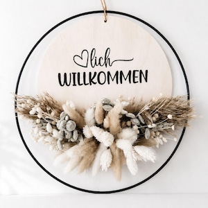 Op de afbeelding: Een ronde houten plank met het Duitse woord "Willkommen" (Welkom) in zwart schrift, geaccentueerd met een hart. Het bord is omlijst door een zwarte metalen ring en versierd met gedroogde bloemen in neutrale tinten.