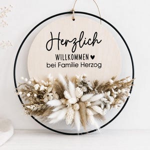 May include: Round wooden sign with a black metal frame, featuring the German phrase "Herzlich Willkommen bei Familie Herzog" in black script. The bottom of the sign is adorned with dried flowers in neutral tones, creating a welcoming aesthetic.