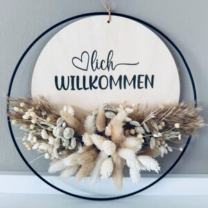 Op de afbeelding: Een ronde houten plank met het woord "Willkommen" in zwart script, geaccentueerd met een hart. Het bord is omgeven door een zwarte metalen ring en versierd met gedroogde bloemen in beige en witte tinten.
