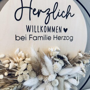 May include: A round wooden sign with the words "Herzlich Willkommen bei Familie Herzog" in black script. The sign is decorated with dried flowers in shades of white, beige, and brown, and is framed by a black metal ring.