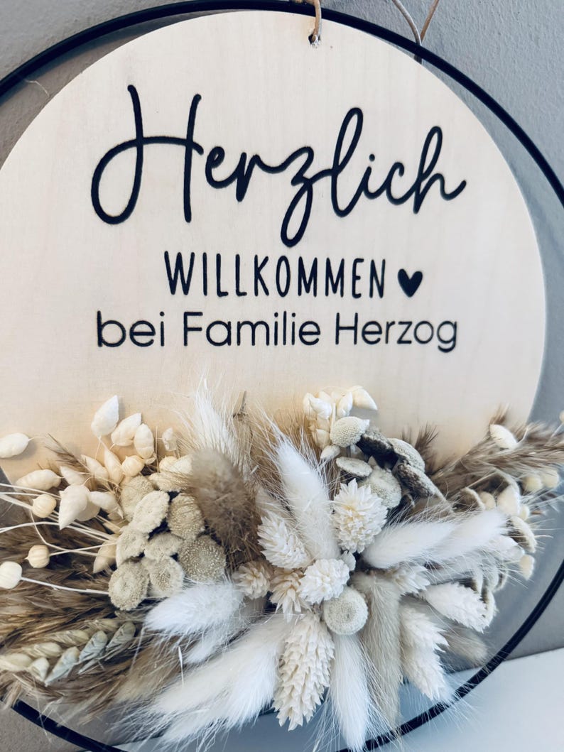 May include: A round wooden sign with the words "Herzlich Willkommen bei Familie Herzog" in black script. The sign is decorated with dried flowers in shades of white, beige, and brown, and is framed by a black metal ring.