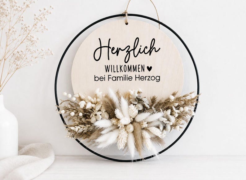 May include: Round wooden sign with a black metal frame, featuring the German phrase "Herzlich Willkommen bei Familie Herzog" in black script. The bottom of the sign is adorned with dried flowers in neutral tones, creating a welcoming aesthetic.