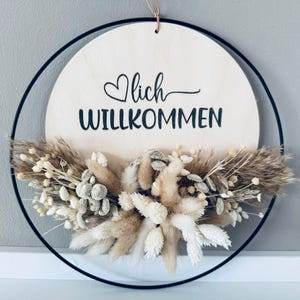 Op de afbeelding: Een ronde houten plank met het woord "Willkommen" in zwart schrift, geaccentueerd met een hart. Het bord is omgeven door een zwarte metalen ring en versierd met gedroogde bloemen in beige en wit. Een decoratief stuk.