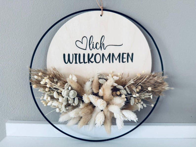 Op de afbeelding: Een ronde houten plank met het woord "Willkommen" in zwart schrift, geaccentueerd met een hart. Het bord is omgeven door een zwarte metalen ring en versierd met gedroogde bloemen in beige en wit. Een decoratief stuk.