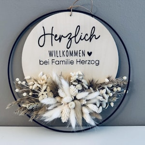 May include: A round wooden sign with the words "Herzlich Willkommen bei Familie Herzog" in black script. The sign is decorated with dried flowers and greenery, and is framed by a black metal ring.