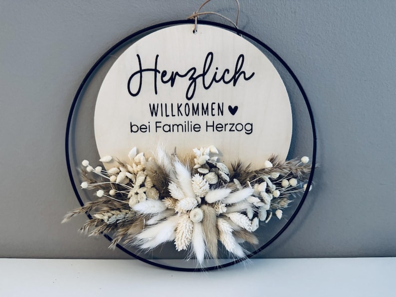 May include: A round wooden sign with the words "Herzlich Willkommen bei Familie Herzog" in black script. The sign is decorated with dried flowers and greenery, and is framed by a black metal ring.