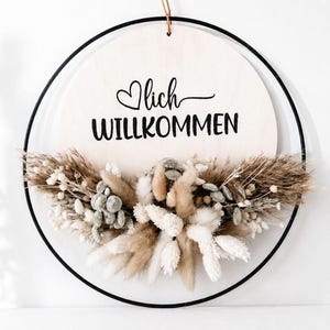 Op de afbeelding: Een ronde houten plank met het woord "Willkommen" in zwart schrift, geaccentueerd met een hart. Het bord is omcirkeld door een zwarte metalen ring en versierd met gedroogde bloemen in wit, beige en bruin. Bovenop zit een natuurlijke touwlus.