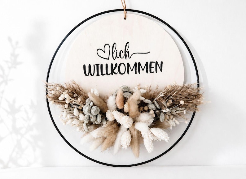 Op de afbeelding: Een ronde houten plank met het woord "Willkommen" in zwart schrift, geaccentueerd met een hart. Het bord is omcirkeld door een zwarte metalen ring en versierd met gedroogde bloemen in wit, beige en bruin. Bovenop zit een natuurlijke touwlus.