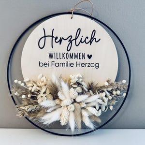 May include: A round wooden sign with the German words "Herzlich Willkommen bei Familie Herzog" in black script. The sign is decorated with dried flowers in shades of white, beige, and brown, and is framed by a black metal ring.