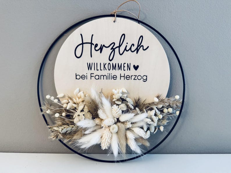 May include: A round wooden sign with the German words "Herzlich Willkommen bei Familie Herzog" in black script. The sign is decorated with dried flowers in shades of white, beige, and brown, and is framed by a black metal ring.