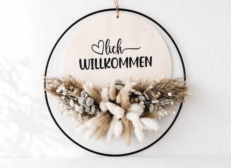 Op de afbeelding: Een ronde houten plank met het Duitse woord "Willkommen" (Welkom) in zwart schrift, geaccentueerd met een hart. Het bord is omlijst door een zwarte metalen ring en versierd met gedroogde bloemen in neutrale tinten.