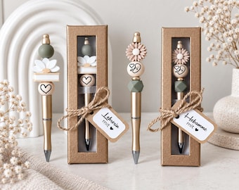 Op de afbeelding: Twee gouden pennen met decoratieve siliconen kralen in olijfgroen, wit en beige. De ene pen heeft een witte bloem en hartkraal, de andere een madeliefje en een baby-vormige kraal. Elke pen zit in een gouden doosje met een label "Lehrerin mit" of "Hebamme mit".