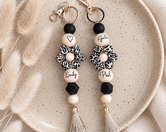 Op de afbeelding: Twee gepersonaliseerde sleutelhangers met zwarte en crèmekleurige kralen, een centrale bloemvormige kraal met een zwart-witte luipaardprint en een beige kwastje. De ene sleutelhanger toont de namen Emily en een hartje, de andere Giulia en Phil.