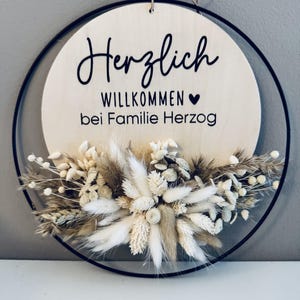 May include: A round wooden sign with the words "Herzlich Willkommen bei Familie Herzog" in black script. The sign is surrounded by a black metal ring and adorned with dried flowers in shades of beige and white. A jute string is attached for hanging.