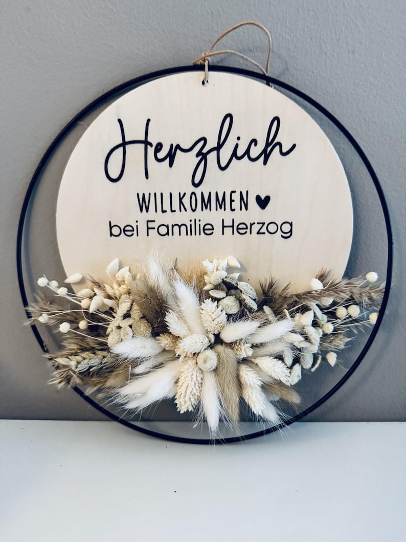 May include: A round wooden sign with the words "Herzlich Willkommen bei Familie Herzog" in black script. The sign is surrounded by a black metal ring and adorned with dried flowers in shades of beige and white. A jute string is attached for hanging.