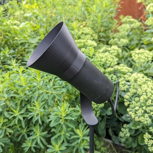 May include: A black outdoor spotlight with a flared head, mounted on a slender black pole. The spotlight is angled towards the viewer, set against a backdrop of green foliage and grass. The spotlight's dimensions are approximately 12 inches in length.