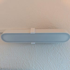 May include: A white and light blue LED light fixture mounted on a textured white wall. The fixture has a rectangular shape with rounded edges and a frosted light diffuser. A white bracket secures the light.