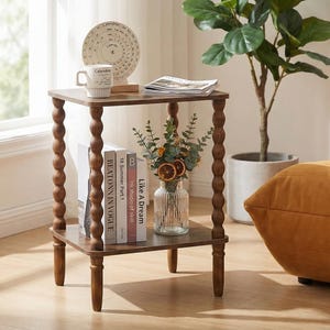 May include: A two-tiered, dark brown wooden side table with turned legs. The top shelf holds a mug, a decorative calendar, and a stack of magazines. The lower shelf displays books and a vase of flowers. A potted plant and a chair are in the background.