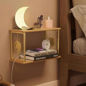May include: A wooden floating shelf with gold accents, featuring a crescent moon lamp, a purple crystal, a pink candle, and a small white clock. The shelf holds books and is mounted on a beige wall, next to a bed.
