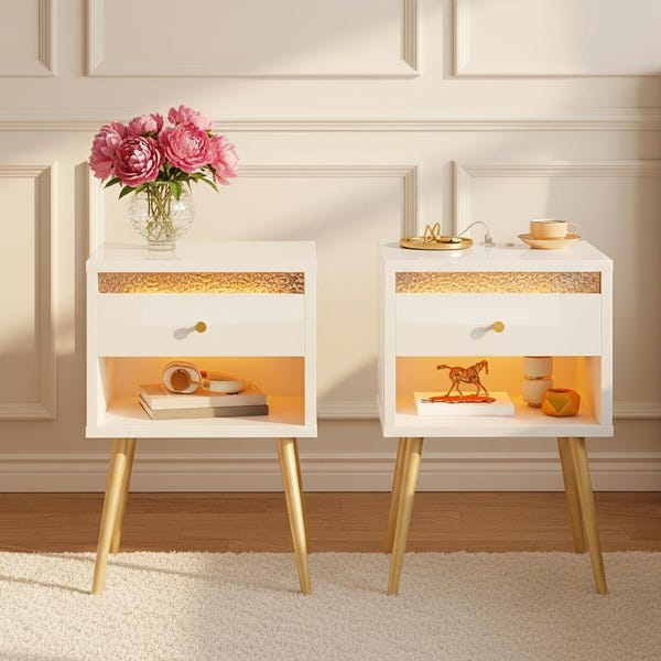 May include: Two white nightstands with gold accents and legs. Each has a drawer with a gold knob and an open shelf. One nightstand has a vase of pink flowers, the other has a decorative horse figurine. A rug and a bed are in the background.