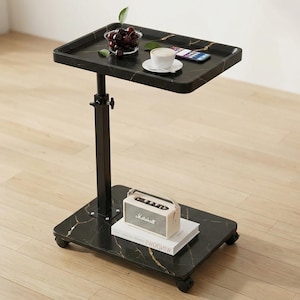 May include: A black marble-patterned adjustable table with a rectangular top and a lower base. The top holds a bowl of cherries, a cup of coffee, and a smartphone. A small speaker rests on the base, along with a stack of books.