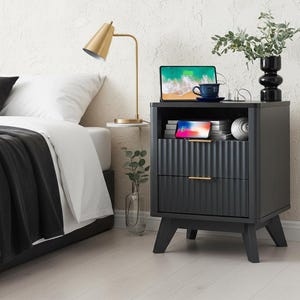 May include: A dark gray nightstand with two drawers and a fluted front. A gold-toned lamp and a tablet sit on top, along with a blue cup and a black vase with greenery. The nightstand is next to a bed with white bedding and a black throw.