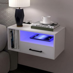 May include: A white floating nightstand with a drawer and open shelf. A lamp, books, and a coffee cup sit on top. The open shelf has a blue LED light and a smartphone. A modern bedroom furniture piece.