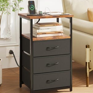 May include: A black and brown nightstand with three drawers. The top shelf holds a phone on a wireless charger, books, and a small clock. The nightstand has a metal frame and is next to a white sofa and a potted plant.