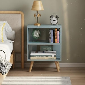 May include: A light blue bedside table with two shelves, holding books, a small green fan, and a clock. A lamp with a beige shade sits on top. The table has wooden legs and is next to a bed with a yellow striped pillow.