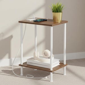 May include: A small, two-tiered side table with a dark brown top and shelf, supported by white legs. A smartphone is charging on the top, next to a small potted plant. White headphones and books rest on the lower shelf.