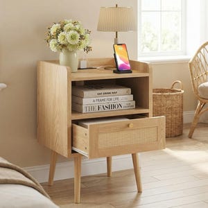 May include: A light wood nightstand with a drawer and open shelf. The drawer has a woven cane front. A lamp, phone, books, and a vase of flowers sit on top. A wicker basket and chair are in the background.