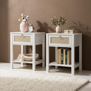 May include: Two white wooden nightstands with a drawer and lower shelf. Each nightstand has a light-colored woven panel on the drawer front. One has a pink vase and a clock, the other has books. The nightstands are on a white rug.