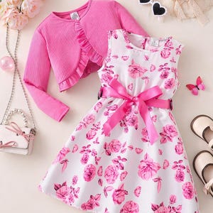 Könnte beinhalten: Ein weißes ärmelloses Kleid mit rosa Blumenmuster und einer passenden rosa Schleife. Eine rosa Bolerojacke mit Rüschenbesatz wird mit dem Kleid kombiniert. Zubehör umfasst eine weiße Handtasche, Sonnenbrille und einen Schmetterling.