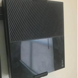 Może przedstawiać: Czarna konsola Xbox zamontowana na białej ścianie. Konsola ma teksturowaną górną część z ukośnymi liniami i gładką, odblaskową dolną część. Logo Xbox jest widoczne w prawym dolnym rogu. Konsola jest utrzymywana na miejscu przez czarne wsporniki.