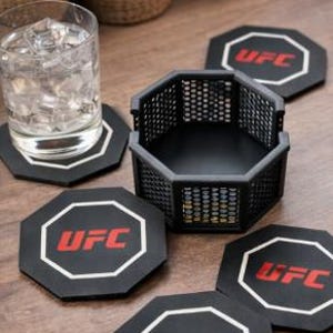 May include: A set of black octagon-shaped coasters with the UFC logo in red and white. A matching black cage-like holder sits nearby, along with a glass of water. The coasters are on a wooden surface.
