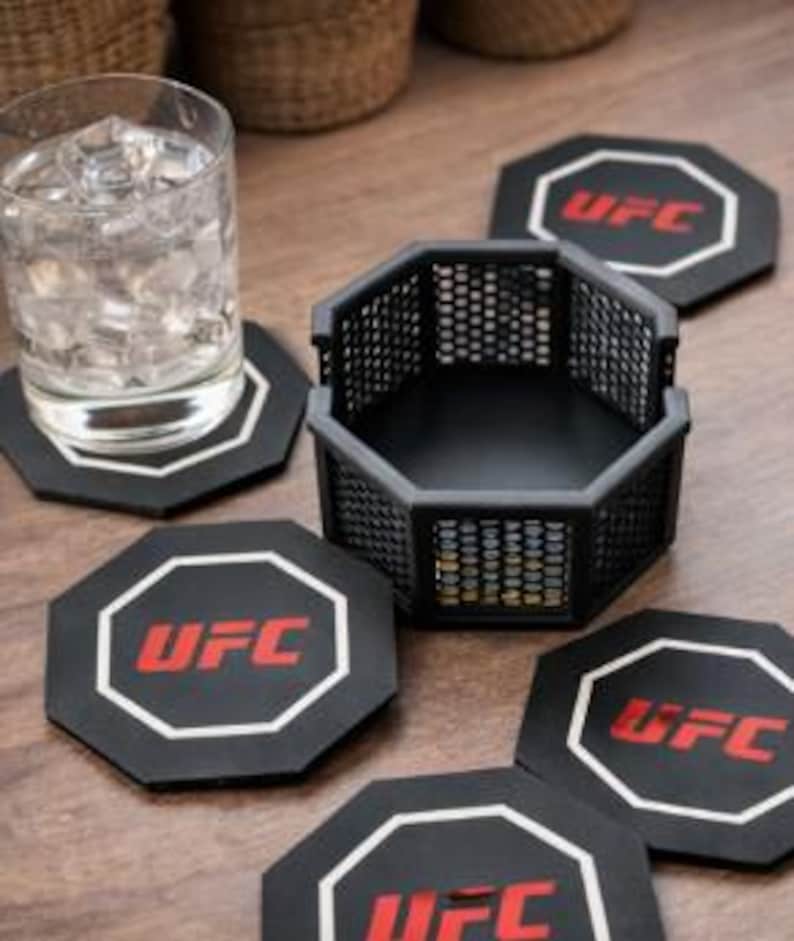 May include: A set of black octagon-shaped coasters with the UFC logo in red and white. A matching black cage-like holder sits nearby, along with a glass of water. The coasters are on a wooden surface.
