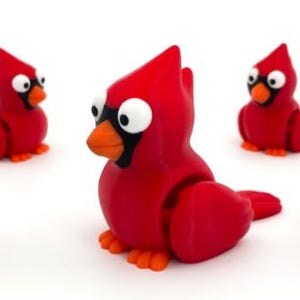May include: A collection of five red cardinal figurines with cartoonish features. Each bird has large, white eyes, a black mask, and an orange beak and feet. The figurines are posed on a white background.