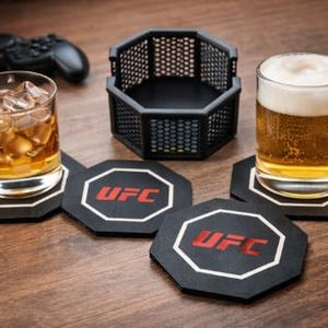 May include: A glass of iced whiskey and a mug of beer sit on black octagonal coasters with the UFC logo. A black cage-like container and a game controller are also visible on a wooden surface.