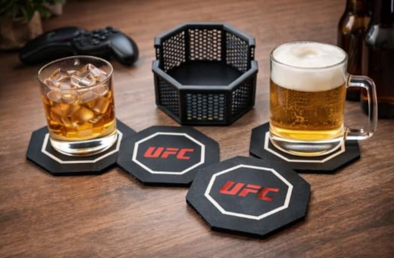 May include: A glass of iced whiskey and a mug of beer sit on black octagonal coasters with the UFC logo. A black cage-like container and a game controller are also visible on a wooden surface.