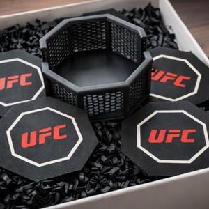 May include: A gift set featuring a black octagon-shaped cage and four matching coasters. Each coaster and the cage have the UFC logo in red and white. The set is presented in a white box with black filler.