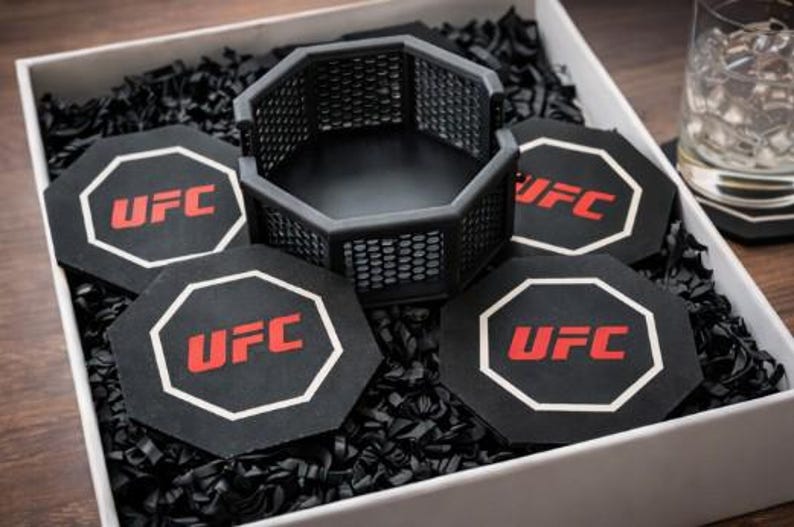 May include: A gift set featuring a black octagon-shaped cage and four matching coasters. Each coaster and the cage have the UFC logo in red and white. The set is presented in a white box with black filler.