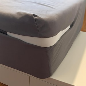 Könnte beinhalten: Ein graues Spannbettlaken bedeckt eine Matratze, wobei eine weiße Seite sichtbar ist. Das Laken ist ordentlich verstaut und zeigt eine saubere, minimalistische Ästhetik. Das Bett steht auf einer weißen Plattform.