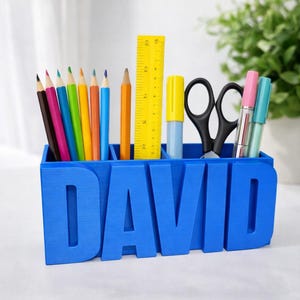 Puede incluir: Un organizador de escritorio azul con el nombre "DAVID" en letras grandes. El organizador contiene lápices de colores, una regla amarilla, un resaltador amarillo, tijeras negras y dos bolígrafos. El organizador está diseñado para guardar artículos de papelería.