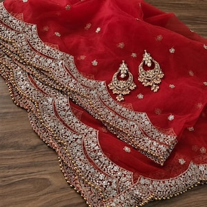 Puede incluir: Un dupatta de tela roja transparente con un intrincado borde festoneado con bordados plateados, lentejuelas y pequeñas cuentas doradas. La tela está adornada con motivos florales dispersos y se acompaña de dos pendientes a juego.