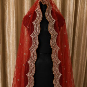 May include: A sheer red veil featuring a scalloped edge with intricate white embroidery and small gold bead detailing along the border. The fabric is adorned with scattered, small white floral motifs throughout the length of the red tulle material.