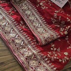 May include: A deep red fabric with intricate gold embroidery and sequin detailing along the border. The textile features a floral pattern and includes a white rectangular tag with the text Tulsie printed in a dark script font with floral accents.